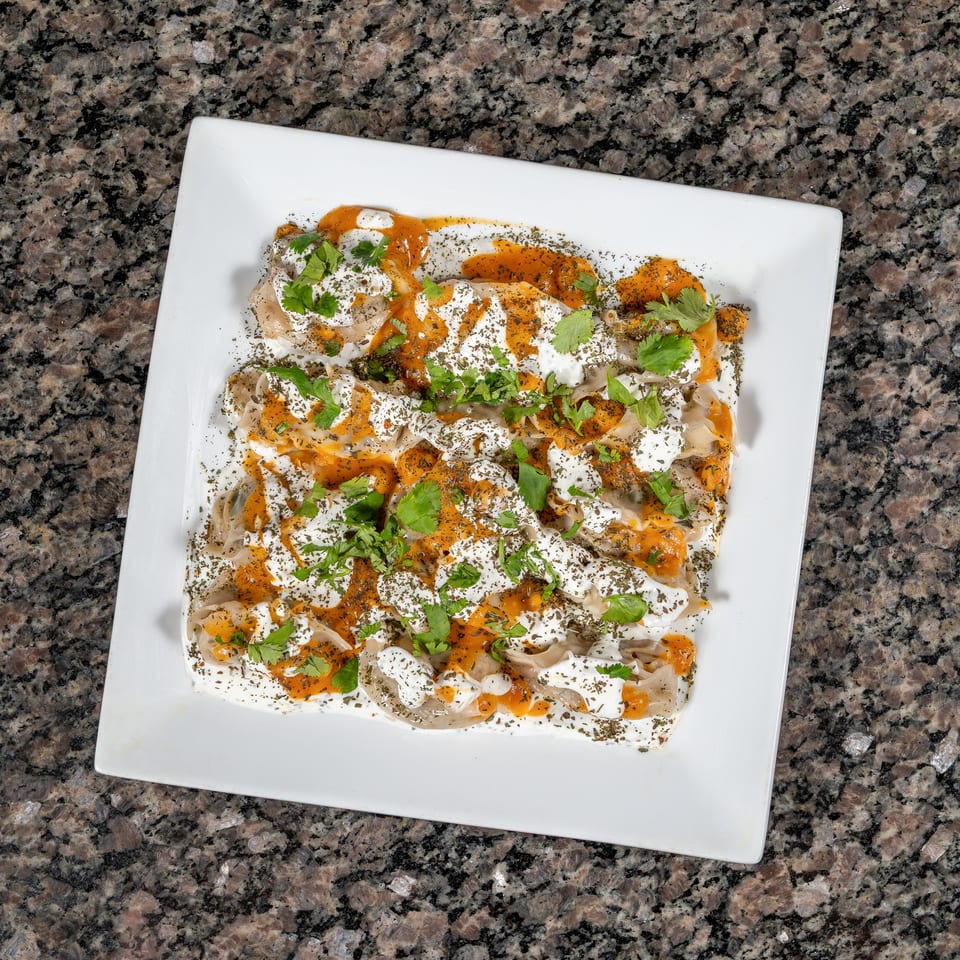 A white rectangular plate filled with Turkish manti, topped with yogurt, red pepper sauce, and herbs.