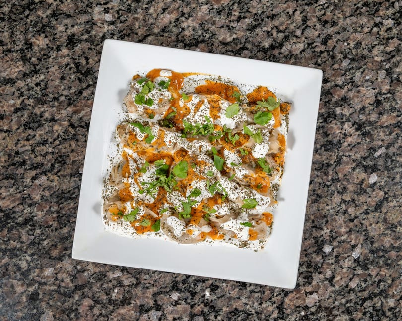 A white rectangular plate filled with Turkish manti, topped with yogurt, red pepper sauce, and herbs.