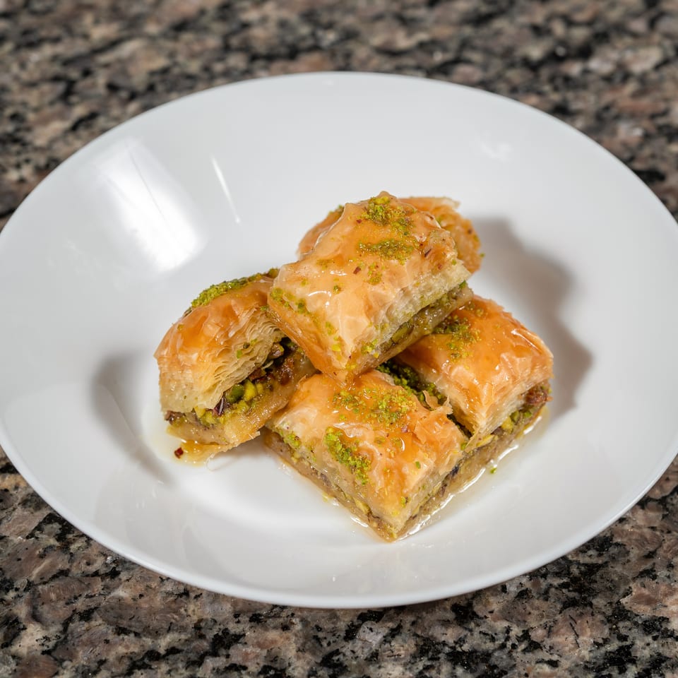 A white plate holds several pieces of golden baklava, drizzled with syrup and sprinkled with pistachios.