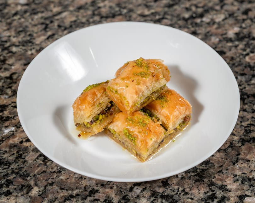 A white plate holds several pieces of golden baklava, drizzled with syrup and sprinkled with pistachios.