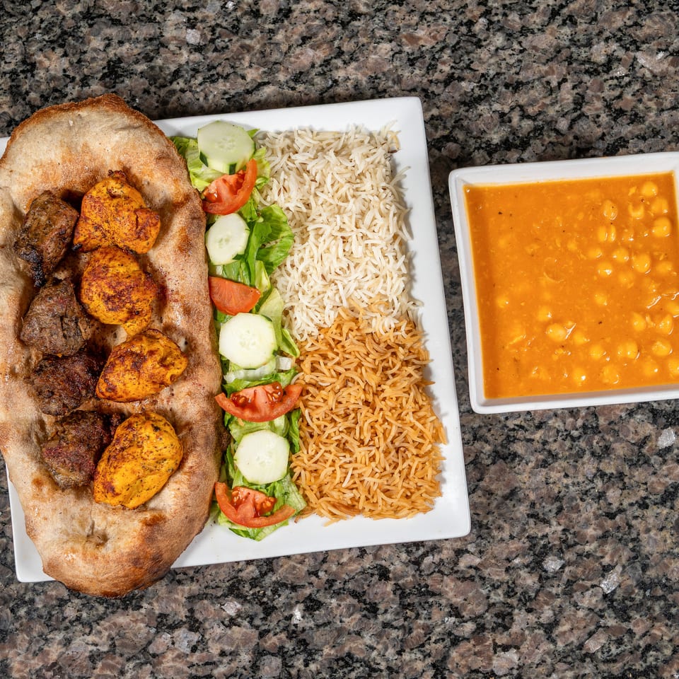Overhead view of a platter with chicken and beef kebabs on naan, rice, salad, and a side of chickpea stew.