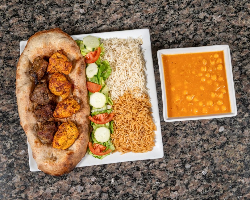 Overhead view of a platter with chicken and beef kebabs on naan, rice, salad, and a side of chickpea stew.