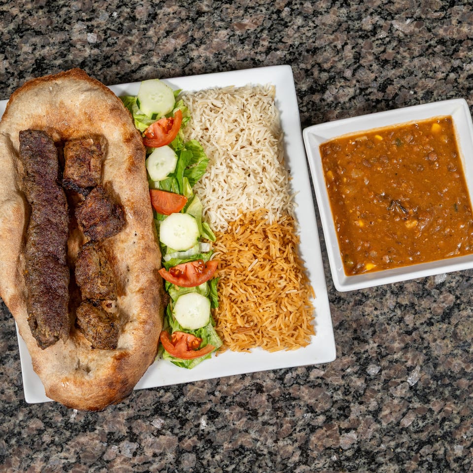 A platter with two types of rice, salad, naan bread with kebabs, and a side of lentil soup.