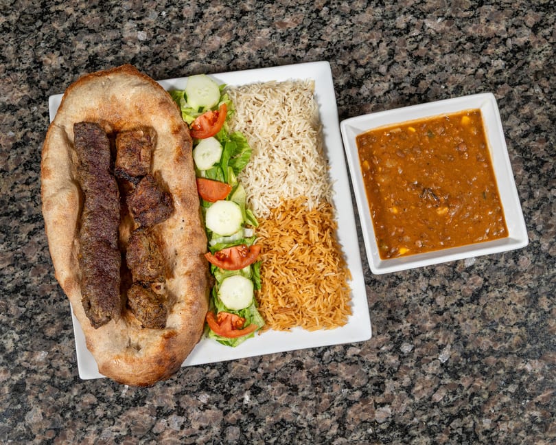 A platter with two types of rice, salad, naan bread with kebabs, and a side of lentil soup.