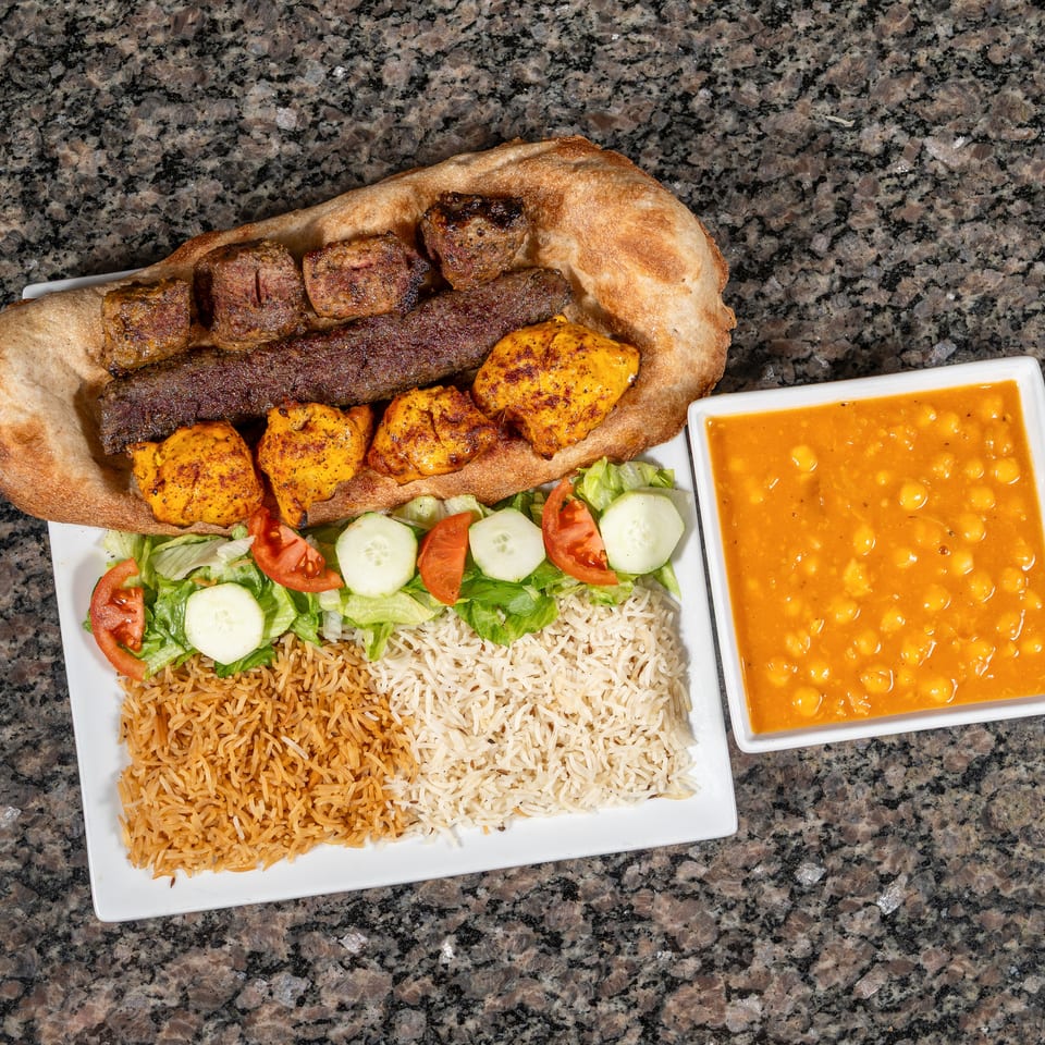 A platter of grilled meats, rice, salad, and a side of chickpea stew on a granite surface.