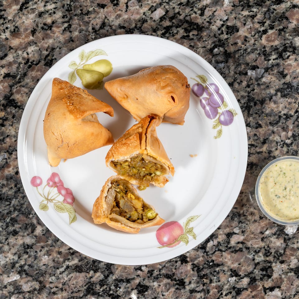 Three golden-brown samosas, one cut open to show a savory filling, on a white plate with dipping sauce.
