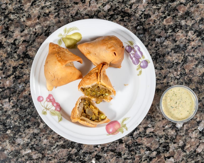 Three golden-brown samosas, one cut open to show a savory filling, on a white plate with dipping sauce.