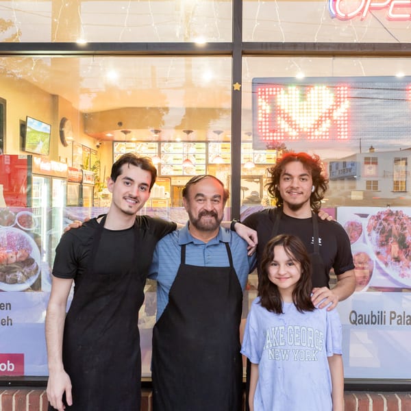 Four smiling people, three men and one child, stand in front of a restaurant window with food posters.